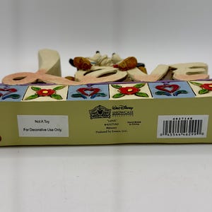 May include: A decorative Disney "Love" figurine. The word "LOVE" is spelled out in large, cream-colored letters, with a cartoon character figure between the "O" and "V". The base is light green with red dots and colorful floral and heart designs.