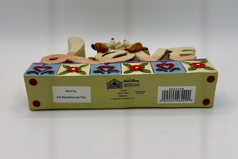 May include: A decorative Disney "Love" figurine. The word "LOVE" is spelled out in large, cream-colored letters, with a cartoon character figure between the "O" and "V". The base is light green with red dots and colorful floral and heart designs.