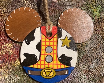 May include: A wooden ornament shaped like Mickey Mouse's head, painted to look like Woody from Toy Story. Woody is wearing his signature cowboy hat, vest, and jeans. The ornament has a brown rope for hanging.