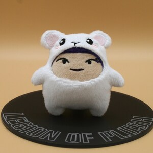 May include: A white plush toy in a bear costume with pink accents, standing on a black circular base. The plush has a smiling face and is set against a light brown background. The base reads "LEGION OF PLUSH".