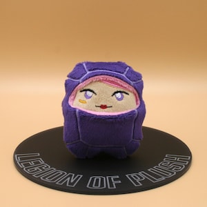 May include: A plush toy with a purple hooded outfit and a face with pink lips, eyes, and a gold earring. The toy is on a black circular base with the text "LEGION OF PLUSH".