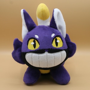 May include: Plush toy of a purple cat-like creature with large yellow eyes, white teeth, and a yellow and white crest. The plush has a smiling expression and is made of soft fabric.