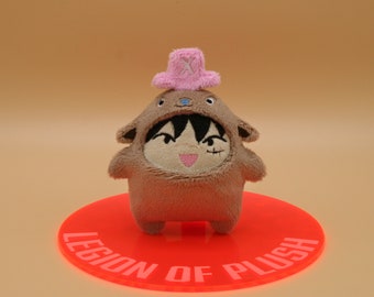 May include: A brown plush toy wearing a pink hat with an 'X' on it. The toy is standing on a red circular base with the text 'Legion of Plush'.