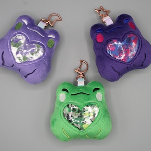 Pode incluir: Três chaveiros de pelúcia em forma de sapo em roxo e verde. Cada chaveiro tem uma janela em forma de coração preenchida com tecido estampado. Eles têm ferragens em tom de ouro rosa e um pequeno laço.