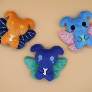 Könnte beinhalten: Drei plüschige, schmetterlingsartige Kreaturen in Orange, Blau und Dunkelblau. Das orangefarbene hat dunkelblaue Flügel, das blaue hat rosa Flügel und das dunkelblaue hat grüne Flügel. Jedes hat einen weißen Bauch und gestickte Details.