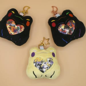 May include: Three plush frog-shaped keychains. Two are black with colorful heart-shaped centers, and the third is yellow with a purple-accented face and a heart-shaped center. Each keychain has a gold or rose gold clasp.