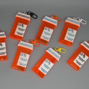 Pode incluir: Chaveiros de frascos de remédios laranja com tampas brancas e fechos de várias cores. Cada chaveiro tem um rótulo branco com texto preto e o símbolo Rx. Alguns frascos contêm pequenas contas laranja.