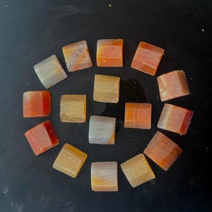 Puede incluir: Una colección de pequeñas gemas cuadradas dispuestas en un patrón circular. Las piedras muestran una variedad de colores, incluyendo naranja, blanco y amarillo, con vetas visibles y texturas naturales. Las gemas probablemente se utilizan para joyería o fines decorativos.