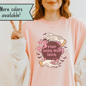 May include: A pink t-shirt with a graphic of a crystal ball with the text "Your baby will latch" inside. The crystal ball is surrounded by bats and two hands with a webbed design.