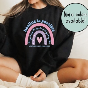 May include: Black sweatshirt with a rainbow graphic and the words "healing is possible." The design includes a heart and the hashtag #EATINGDISORDERAWARENESS. The sweatshirt is a crew neck style.