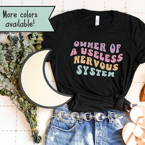 May include: Black t-shirt with the text "OWNER OF A USELESS NERVOUS SYSTEM" in rainbow colors. The shirt is worn with blue jeans and a belt with star-shaped buckles.