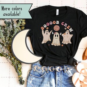 May include: Black t-shirt featuring the phrase "BOOOOB CREW" above three cartoon ghosts. The ghosts are in shades of beige and white with floral accents. The shirt has a crew neck and short sleeves. The words "More colors available!" are in a green banner.