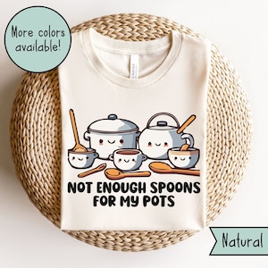 Pode incluir: Uma camiseta branca com um gráfico de panelas e colheres de desenhos animados. O texto na camiseta diz "Não há colheres suficientes para minhas panelas".