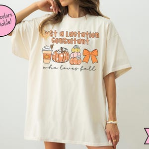 May include: Ivory-colored t-shirt with the text "Just a Lactation Consultant who loves fall." The shirt features fall-themed graphics, including a pumpkin spice latte, pumpkins, and a bow.