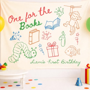 May include: A cream-coloured backdrop with colourful illustrations and text for a birthday party. The text reads "One for the Books" and "Liam's First Birthday." Balloons and a small table with cupcakes are also visible.