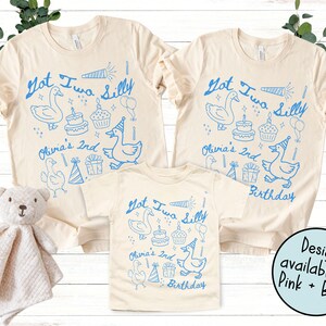 May include: Three beige t-shirts with blue graphics. The shirts feature the text "Got Two Silly" and "Olivia's 2nd Birthday" with illustrations of a goose, cake, cupcake, and party hat. A teddy bear is in the image.