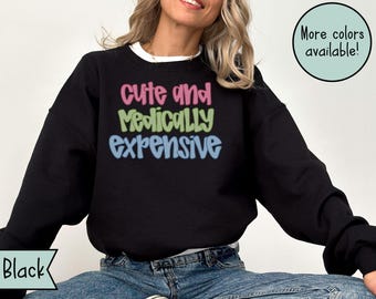 May include: Black sweatshirt with the text "cute and medically expensive" in colorful bubble letters. The sweatshirt has a crew neck and long sleeves. The words "More colors available!" are in a green circle.