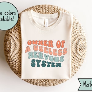 May include: A white t-shirt with the text "OWNER OF A USELESS NERVOUS SYSTEM" in a retro, colorful font. The text is in shades of blue, green, orange, and pink.