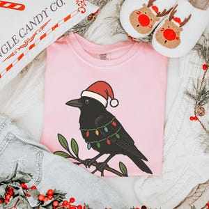 May include: A light pink t-shirt featuring a black crow wearing a Santa hat and Christmas lights. The crow is perched on a branch with green leaves. The image also includes a pillow with "KRINGLE CANDY CO." text, reindeer slippers, and denim.