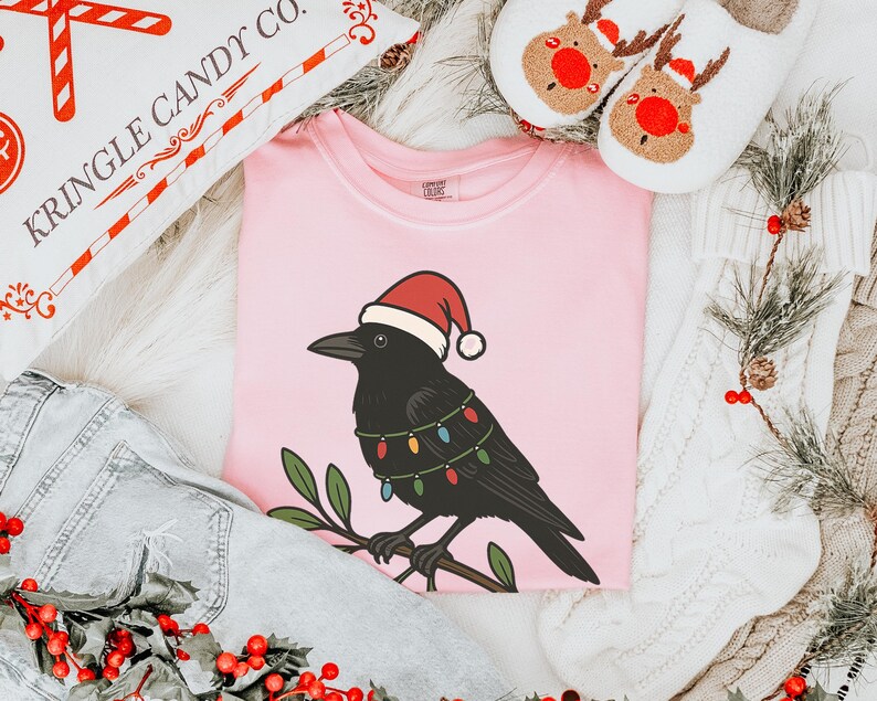 May include: A light pink t-shirt featuring a black crow wearing a Santa hat and Christmas lights. The crow is perched on a branch with green leaves. The image also includes a pillow with "KRINGLE CANDY CO." text, reindeer slippers, and denim.