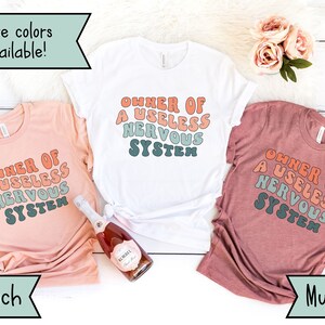 May include: Three t-shirts in peach, white, and mauve colors with the text "OWNER OF A USELESS NERVOUS SYSTEM" printed in a retro font style.