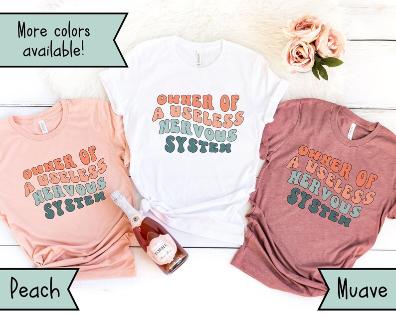 May include: Three t-shirts in peach, white, and mauve colors with the text "OWNER OF A USELESS NERVOUS SYSTEM" printed in a retro font style.