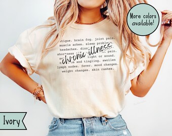 May include: Ivory-colored t-shirt with the words "chronic illness" in a handwritten font. The shirt lists symptoms such as fatigue, brain fog, joint pain, and skin rashes. A teal circle with the text "More colors available!" is in the upper right corner.