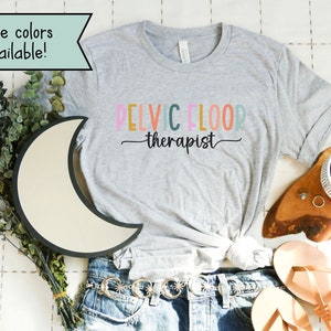 May include: A gray t-shirt with the text "PELVIC FLOOR therapist" in rainbow colors. The shirt is on a white surface with a moon-shaped light, a wooden tray with a coffee cup, and a pair of sandals.