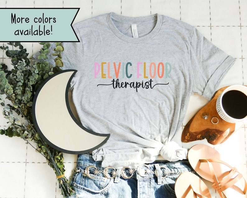 May include: A gray t-shirt with the text "PELVIC FLOOR therapist" in rainbow colors. The shirt is on a white surface with a moon-shaped light, a wooden tray with a coffee cup, and a pair of sandals.