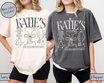 May include: Two t-shirts in ivory and pepper colors with the text "Katie's" and a graphic of a cocktail glass, a bride's veil, and a party hat. The text below the graphic says "Here comes the bride/party Bachelorette Party".