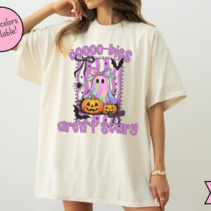 May include: Ivory t-shirt with a Halloween graphic. The design features a ghost, pumpkins, and bats, with the text "Boo-bies aren't scary" in purple and black. The shirt has a loose fit.