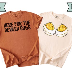 Pode incluir: Duas camisetas, uma cor inhame e outra marfim. A camiseta cor inhame tem o texto preto "HERE FOR THE DEVILED EGGS". A camiseta marfim apresenta um gráfico de dois ovos recheados com recheio amarelo.