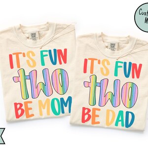 May include: Two ivory-colored t-shirts with the text "IT'S FUN TWO BE MOM" and "IT'S FUN TWO BE DAD" in rainbow colors. Each shirt has a Comfort Colors tag and a gold necklace. The shirts are a matching set.
