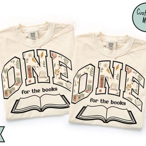 May include: Two ivory-colored t-shirts with the word "ONE" in a black, floral-filled design, and the text "for the books" below an open book illustration. A gold necklace is visible on each shirt. The shirts have a round neckline.