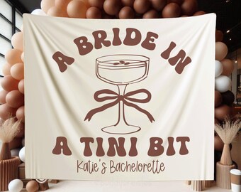 Könnte beinhalten: Ein weißes Banner mit braunem Text, der "A Bride In A Tini Bit" lautet, mit einer stilisierten Illustration eines Cocktailglases. Das Banner enthält auch den Text "Katie's Bachelorette".