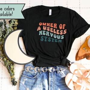 May include: Black t-shirt with a retro-style graphic that reads "Owner of a Useless Nervous System". The shirt is laid out on a white surface with a pair of sandals, a belt, and a cup of coffee.
