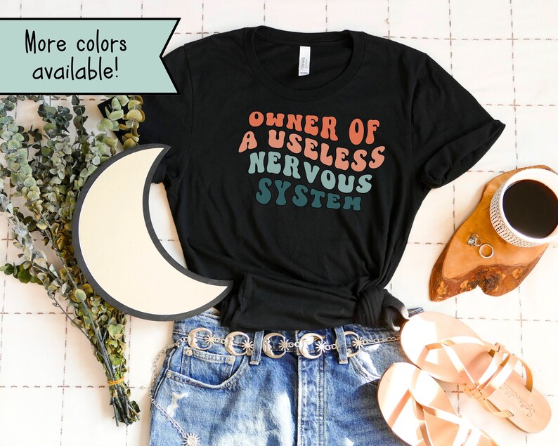 May include: Black t-shirt with a retro-style graphic that reads "Owner of a Useless Nervous System". The shirt is laid out on a white surface with a pair of sandals, a belt, and a cup of coffee.