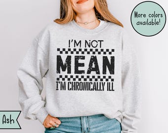 May include: A light gray sweatshirt with the text "I'M NOT MEAN I'M CHRONICALLY ILL" in black block letters. The text is framed by a checkered pattern. The words "More colors available!" are in a green circle.