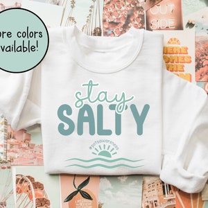 May include: White sweatshirt with the text "stay SALTY" in teal blue lettering. The text is styled with a wavy font. Below the text is a teal blue sun with waves and the hashtag #potsawareness.