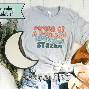 May include: A gray t-shirt with the text "OWNER OF A USELESS NERVOUS SYSTEM" in a rainbow color scheme. The text is in a retro font style.