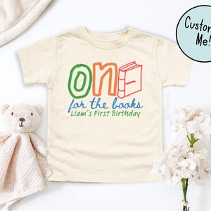 May include: A cream-colored toddler t-shirt with the text "One for the books" in green, orange, and red. The text "Liam's First Birthday" is written in blue. A stuffed teddy bear and a blanket are on the left side of the shirt.