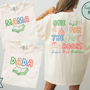 Puede incluir: Camisetas de color marfil con texto y gráficos coloridos. Una camiseta dice "MAMA" encima de un gráfico de un libro abierto. Otra dice "DADA" encima de un libro. Una tercera camiseta tiene "ONE FOR THE BOOKS" con gráficos temáticos de cumpleaños y "Liam's First Birthday".
