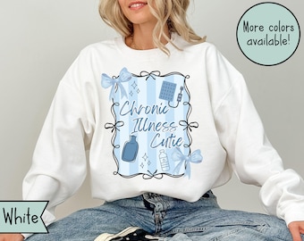 May include: White sweatshirt with a blue and white graphic that reads "Chronic Illness Cutie." The graphic includes blue bows, a bottle, and other medical-themed illustrations. The text "More colors available!" is in a green circle.