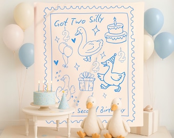 Puede incluir: Un telón de fondo rosa claro con dibujos azules de gansos, globos y una tarta, con el texto "Got Two Silly Second Birthday". Una tarta azul claro con velas está sobre una pequeña mesa blanca. Dos juguetes de peluche de gansos están en primer plano, con globos en el fondo.
