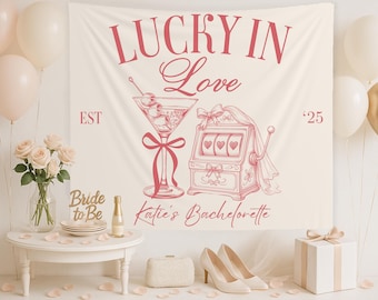 May include: A cream-colored banner with the text "LUCKY IN Love" and "Katie's Bachelorette" in pink script. The banner features illustrations of a martini glass and a slot machine. The scene includes balloons, flowers, and a table with shoes.