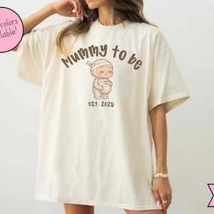 May include: Ivory-colored t-shirt with the text "Mummy to be" and a cartoon mummy graphic. The graphic includes the text "est. 2025". A pink circle with the text "More colors available!" is in the upper left corner.