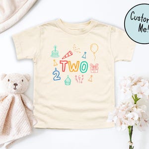 May include: Cream-colored toddler t-shirt with colorful birthday-themed graphics. The word "TWO" is displayed in rainbow colors, surrounded by illustrations of a cake, party hat, balloon, and gift. A "Customize Me!" button is in the upper right.