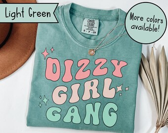 Op de afbeelding: Een lichtgroen T-shirt met de tekst "DIZZY GIRL GANG" in roze, perzikkleurige en groene letters. De letters zijn in een retro, bubbelvormig lettertype. Er zijn kleine witte sterren rond de tekst.