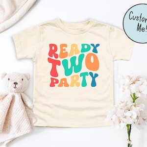 Puede incluir: Camiseta crema para niños pequeños con las palabras "READY TWO PARTY" en letras coloridas de estilo retro. Un osito de peluche y una manta suave están a la izquierda, y un ramo de flores blancas a la derecha. Un círculo azul con el texto "Customize Me!" está en la parte superior derecha.
