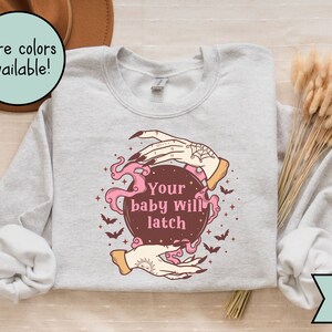 May include: A light gray crewneck sweatshirt featuring a graphic of two hands holding a dark brown crystal ball with pink smoke, stars, and bats. The text "Your baby will latch" is printed in pink in the center of the crystal ball.